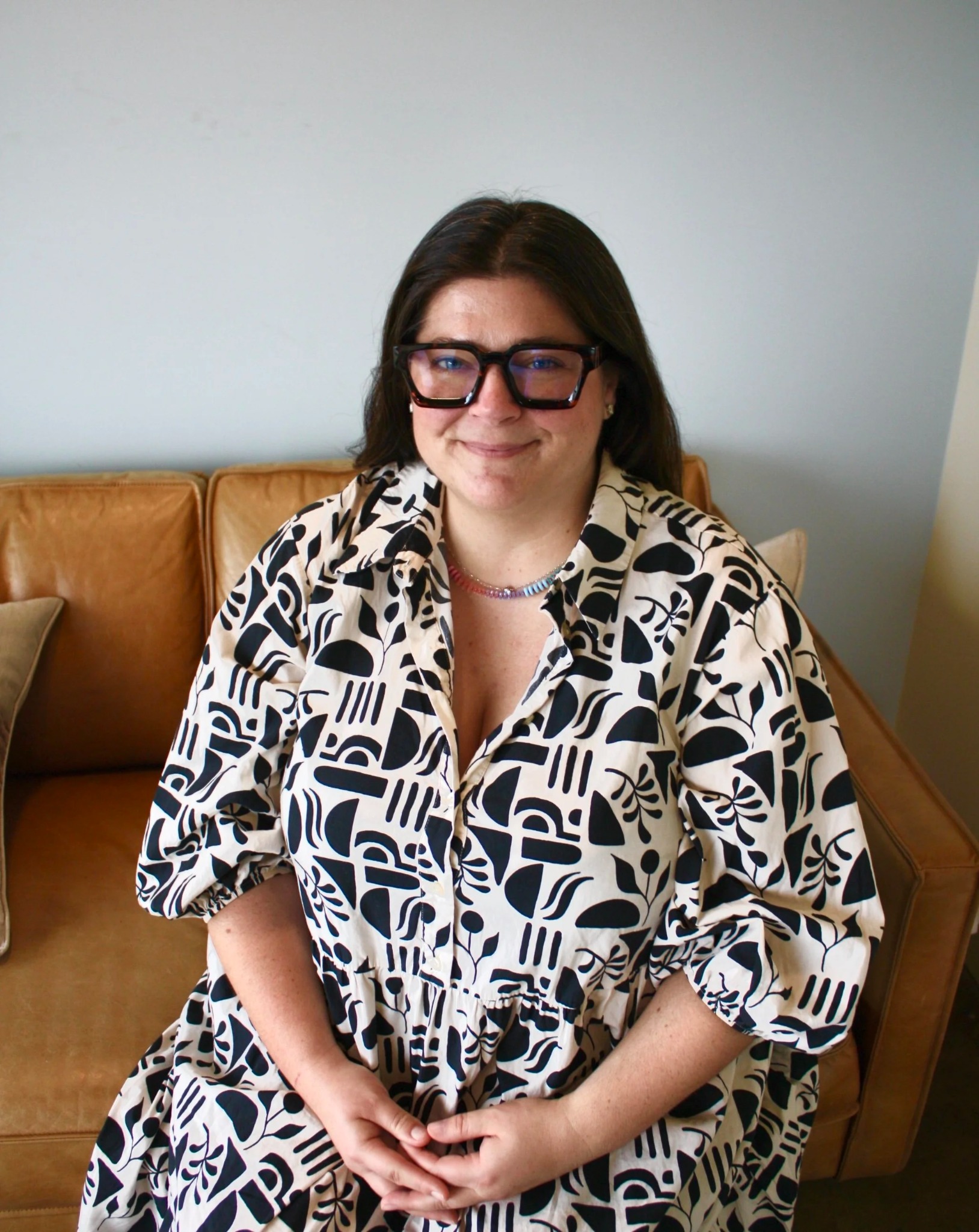 Laura Magliochetti smiling on a tan sofa, wearing black glasses and a black-and-white patterned outfit